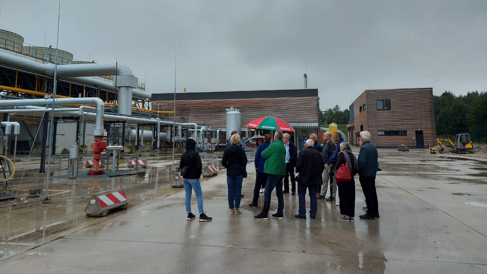 Eine Gruppe von Menschen steht auf einem nassen Industriegelände mit Rohren, Gebäuden und Maschinen - Teil der Bemühungen, Wärmenetze auszubauen. Einige tragen Jacken und haben Regenschirme in der Hand. Der Himmel ist bedeckt und der Boden glänzt vom Regen.