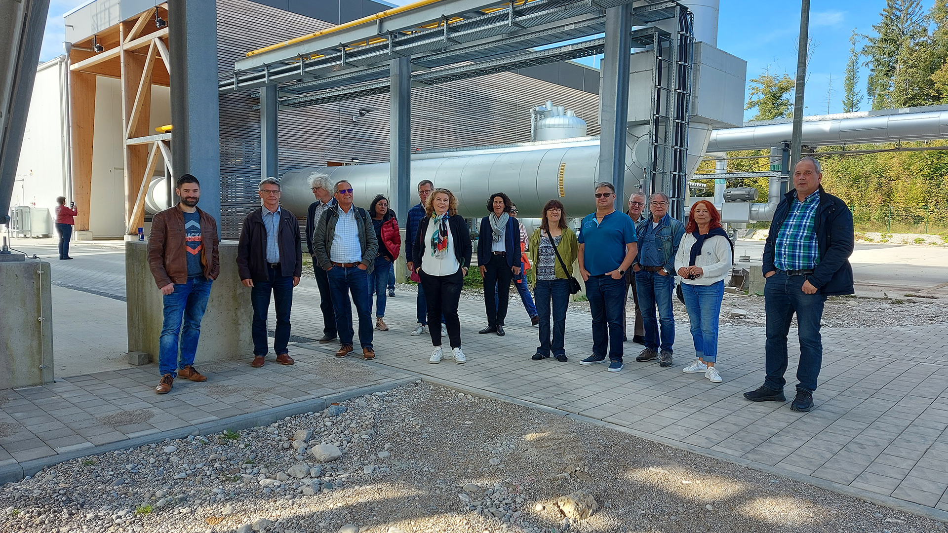Eine Gruppe von dreizehn Erwachsenen steht an einem sonnigen Tag auf einer gepflasterten Fläche im Freien in der Nähe von Industrierohren und -anlagen und posiert für ein Foto. Im Hintergrund sind Bäume und Industriegebäude im Zusammenhang mit der deutschen Erdwärme und der Tiefengeothermie zu sehen.