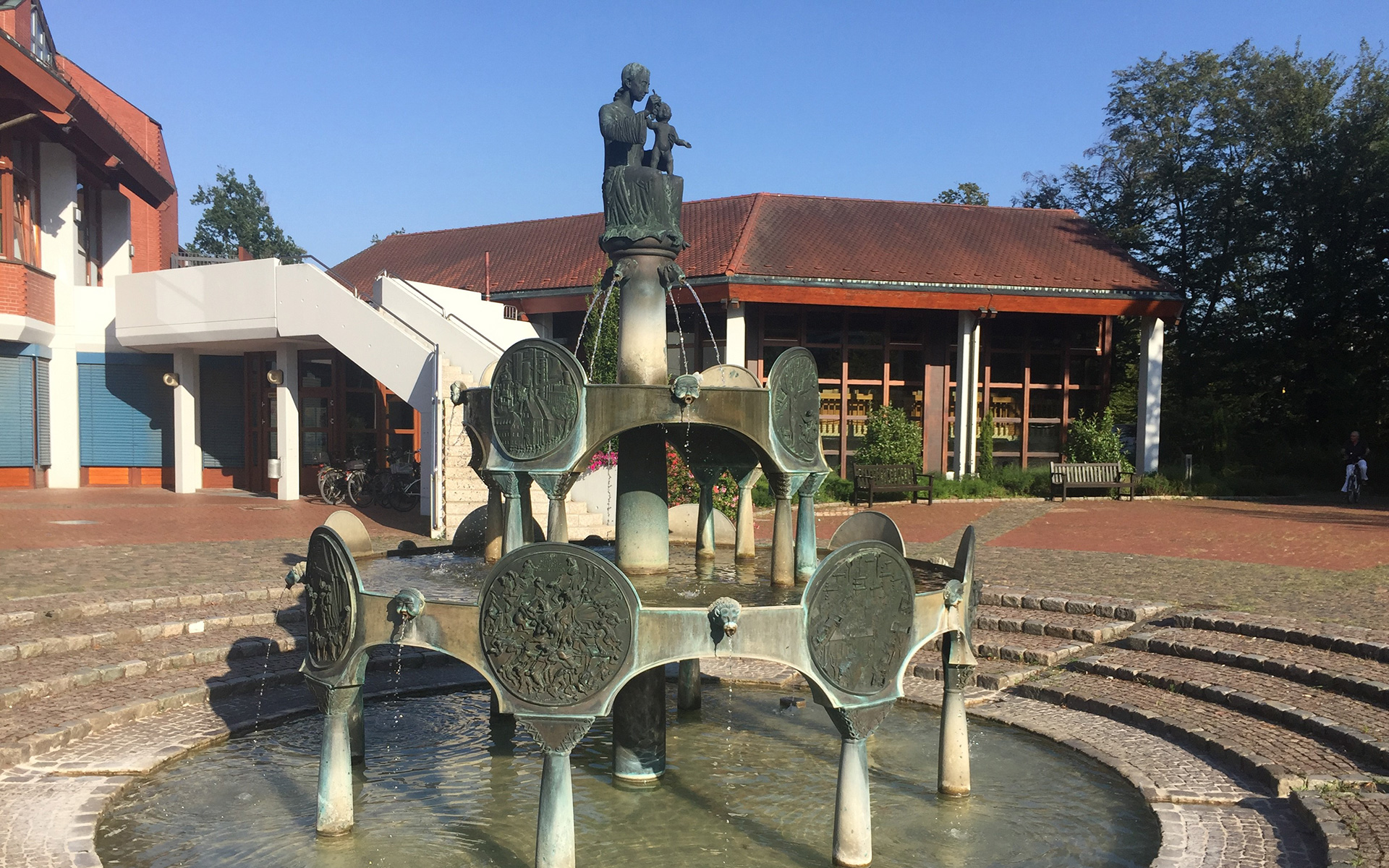 Ein dekorativer Bronzebrunnen mit kreisrunden Tafeln steht auf einem gepflasterten Platz vor einem rotgedeckten Gebäude, das Innovation wie Tiefengeothermie für Dekarbonisierung symbolisiert, umgeben von Bäumen unter einem klaren blauen Himmel.