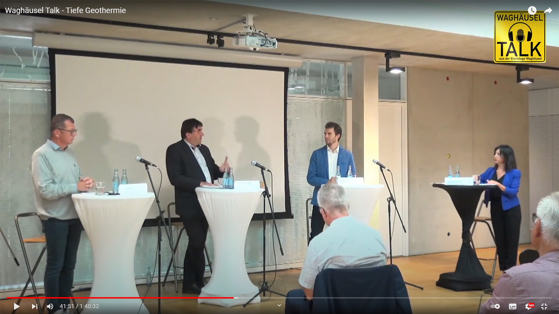 Vier Personen diskutieren auf einer Podiumsbühne mit weißen Podien und Mikrofonen über Themen wie Dekarbonisierung und Wärmeversorgnung. Drei Männer und eine Frau stehen sich gegenüber, während das Publikum zuhört, mit einem gelben Waghäusel Talk-Schild in der oberen rechten Ecke.