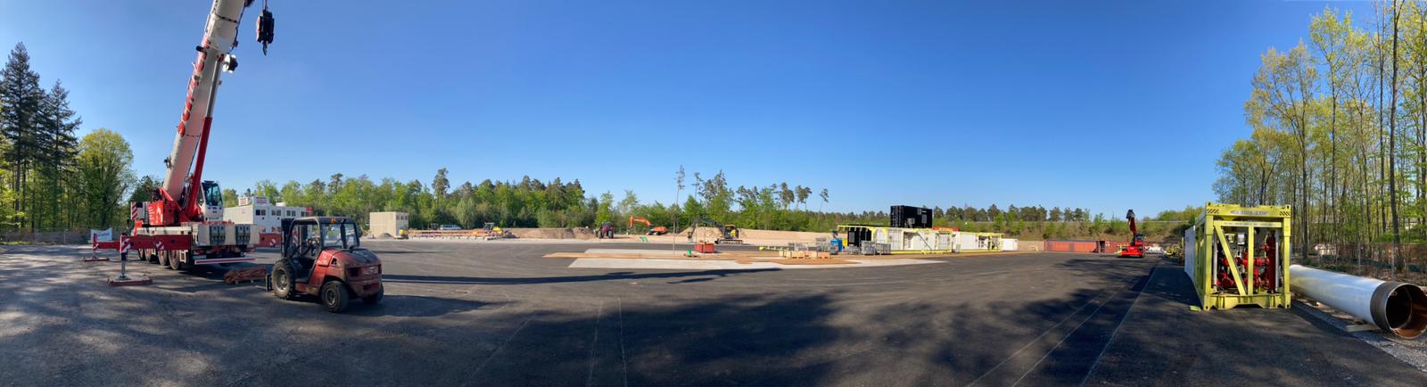 Weite Sicht auf eine Baustelle mit Schwerpunkt Wärmeversorgung, mit schweren Maschinen wie einem Kran und einem Gabelstapler zwischen verstreuten Materialien. Unfertige Bauwerke stehen zwischen Bäumen und blauem Himmel und verdeutlichen die Bemühungen der deutschen Erdwärme.
