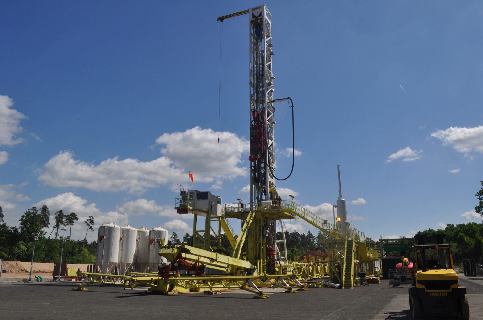 Ein großer gelber Bohrturm steht auf einem gepflasterten Gelände unter einem blauen Himmel mit vereinzelten Wolken. Im Hintergrund sind Bäume und weiße Lagertanks zu sehen, während deutsche Erdwärme Wärmenetze mit Maschinen und Arbeitern in der Nähe ausbaut.