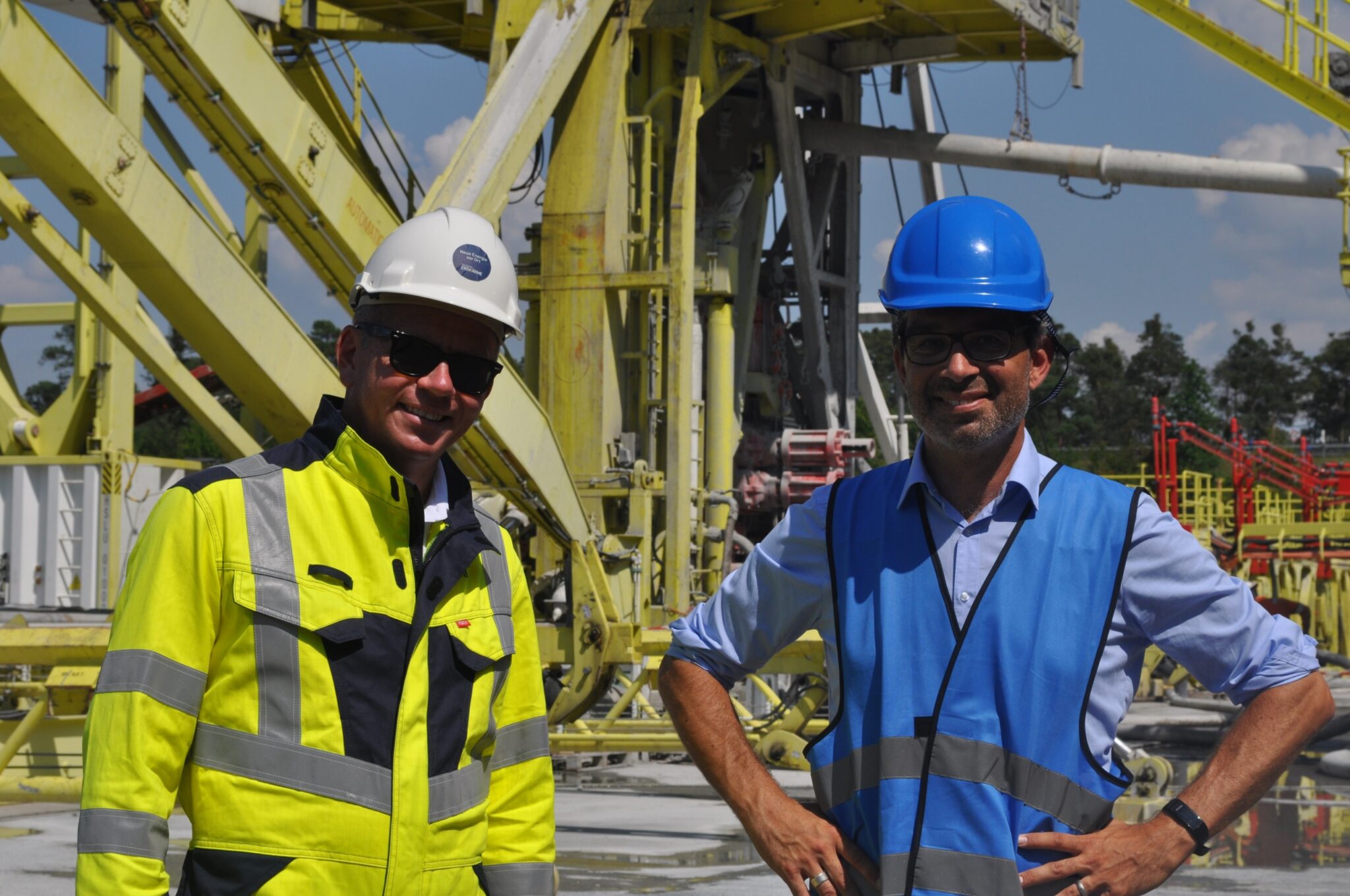 Zwei Männer mit Sicherheitshelmen und Warnkleidung stehen auf einem Industriegelände mit großen gelben Maschinen und Rohren und diskutieren darüber, wie man Wärmenetze für eine effizientere Wärmeversorgung ausbaut. Der eine trägt eine gelbe Jacke, der andere eine blaue Weste.