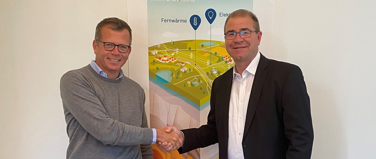 Zwei Männer in Geschäftskleidung lächeln und schütteln sich die Hände vor einem farbenfrohen, illustrierten Plakat, das eine Landschaft mit beschrifteten Abschnitten Fernwärme und Elektro zeigt und auf die Bemühungen hinweist, Wärmenetze für eine sichere Wärmeversorgung auszubauen.