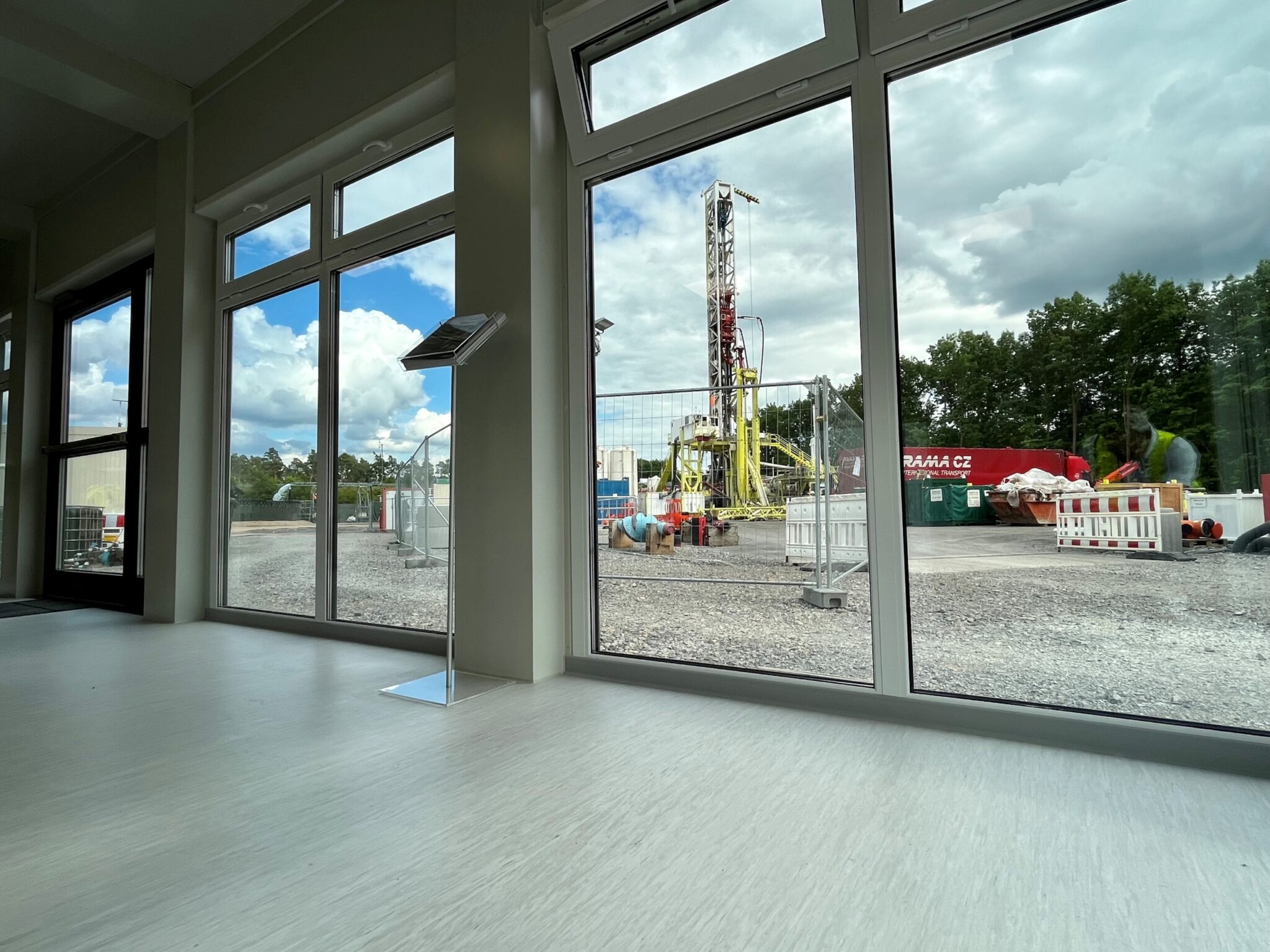 Blick aus dem Inneren eines Gebäudes durch große Fenster auf eine Bohrstelle der deutschen Erdwärme, mit einem hohen Bohrturm, Ausrüstung und Bäumen im Hintergrund unter einem teilweise bewölkten Himmel.