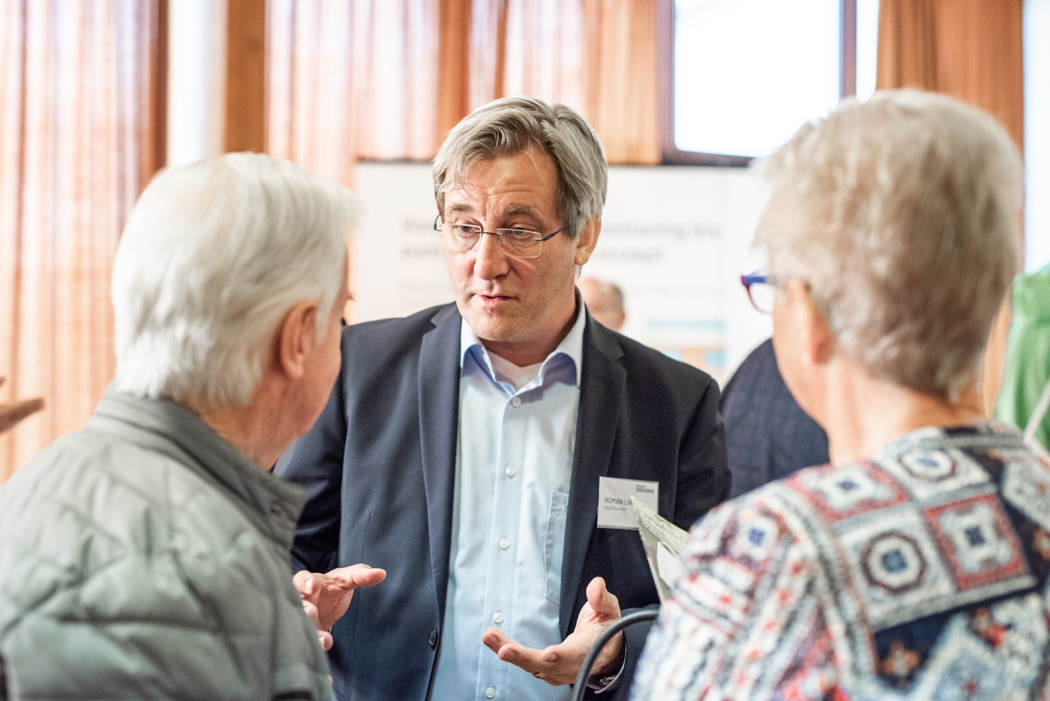 Ein Mann im Anzug und mit Brille spricht mit zwei älteren Erwachsenen bei einer Indoor-Veranstaltung über deutsche Erdwärme. Er gestikuliert mit seinen Händen, während sie zuhören. Der Hintergrund zeigt unscharfe Menschen und Fenster mit Vorhängen.