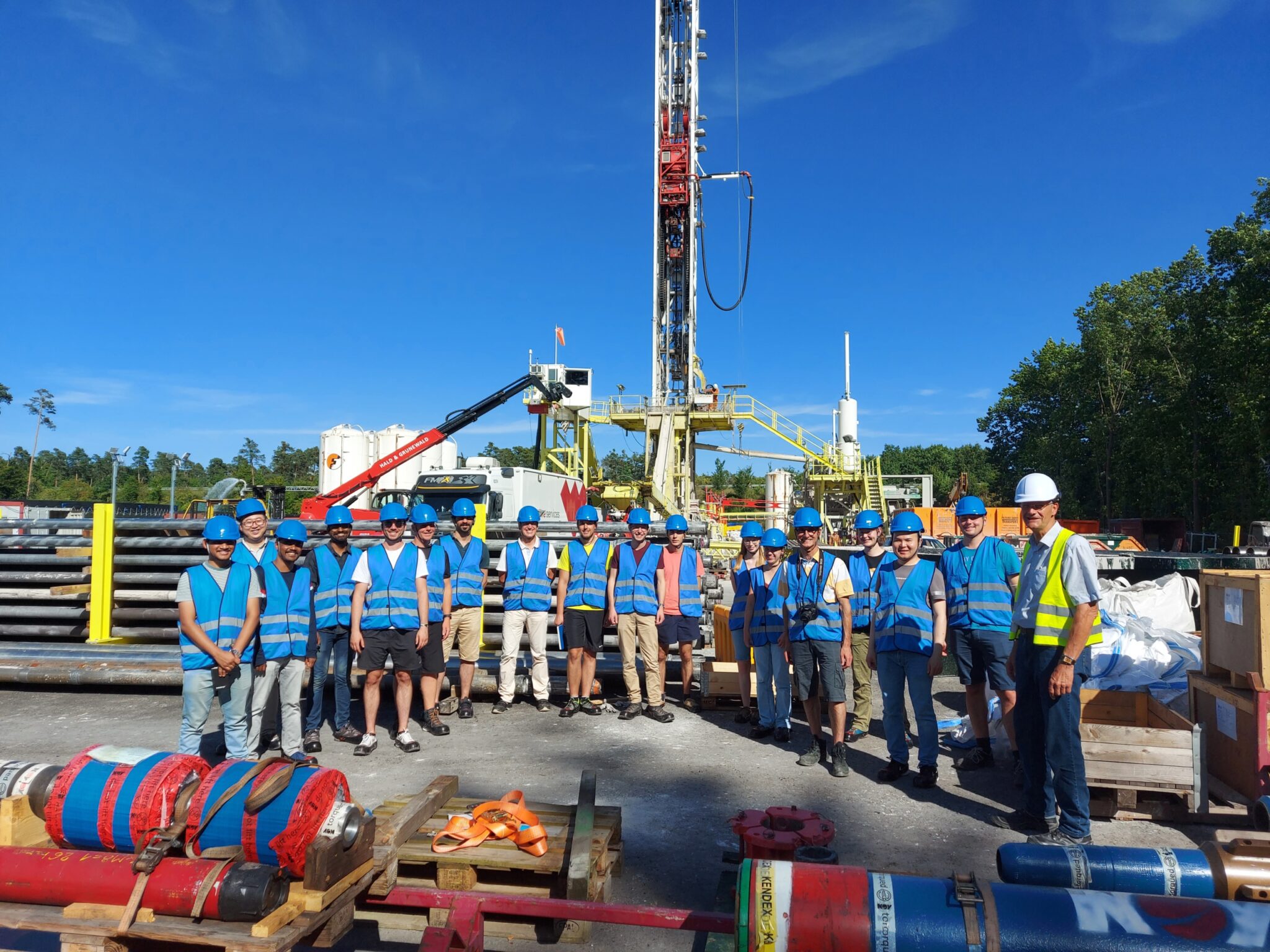 Eine Gruppe von Arbeitern in blauen Sicherheitswesten und Helmen steht auf einem Außengelände der Tiefengeothermie-Bohranlage der deutschen Erdwärme zusammen, umgeben von Geräten und Rohren unter einem klaren blauen Himmel.