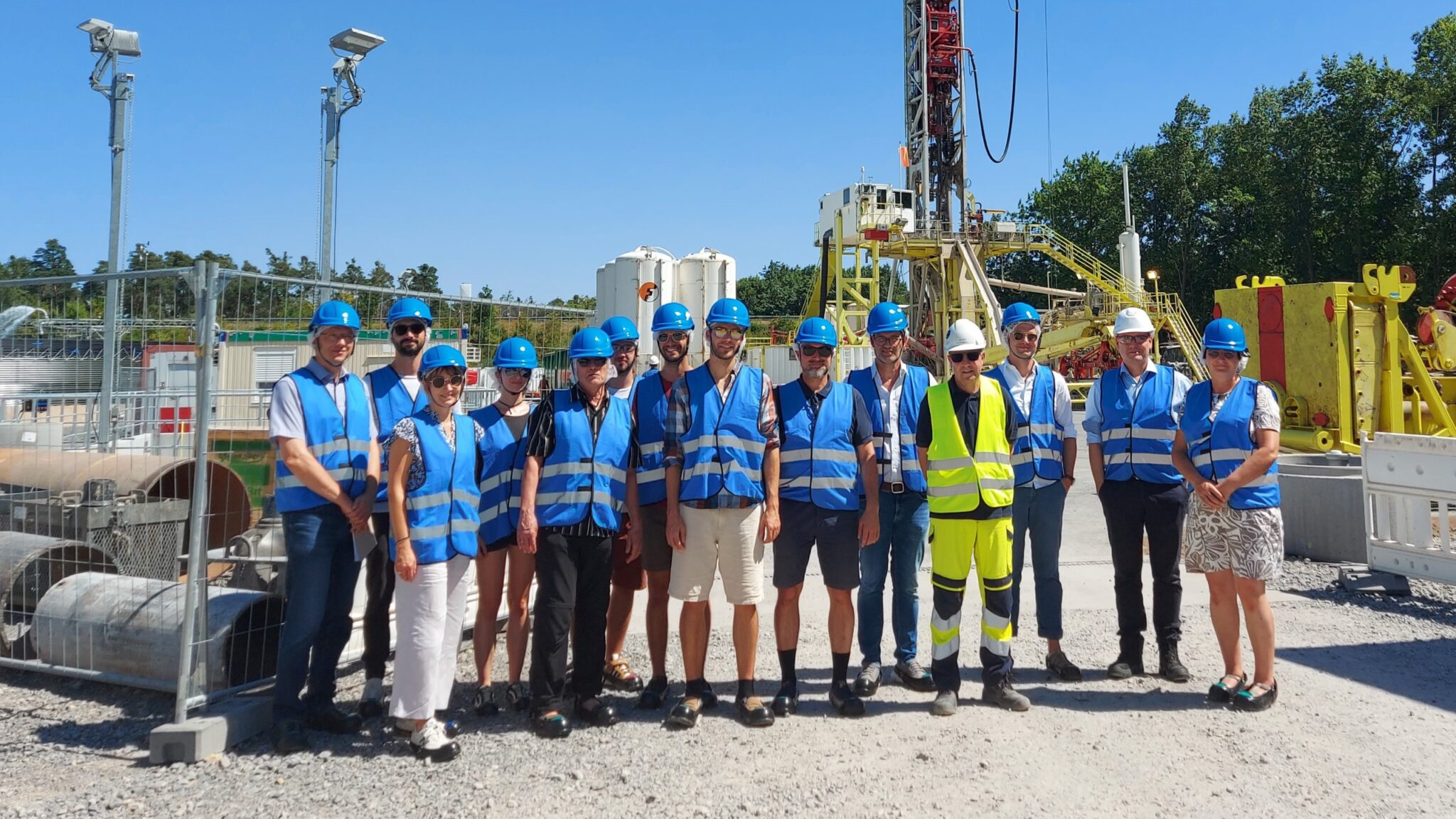 Eine Gruppe von Personen mit blauen Sicherheitswesten und Schutzhelmen steht an einem sonnigen Tag auf einem Industriegelände im Freien zusammen und arbeitet an der Dekarbonisierung und Wärmeversorgnung durch das Ausbreiten von Wärmenetzen in der Nähe von Maschinen.