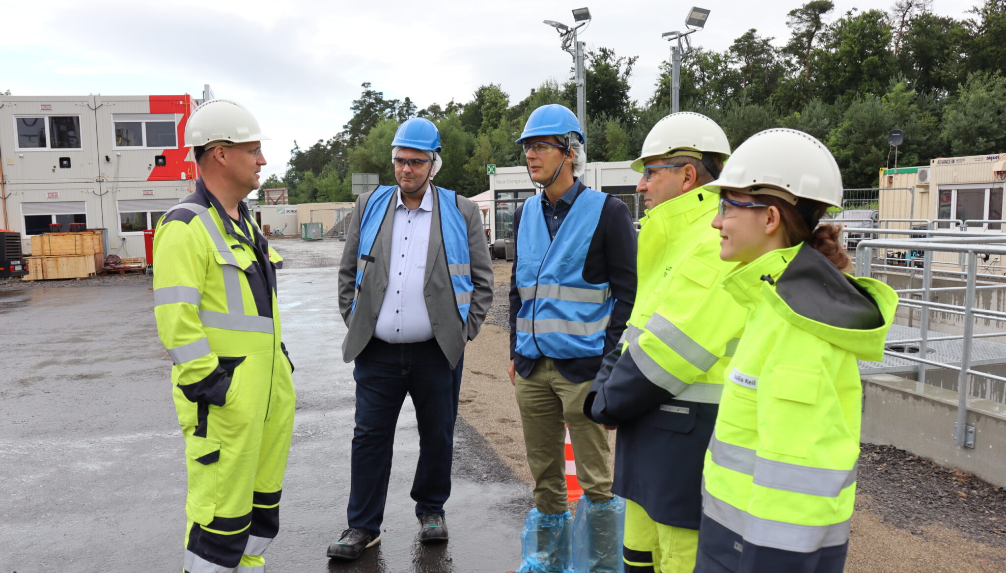 Eine Gruppe von fünf Personen mit Sicherheitsausrüstung, darunter Helme und Warnwesten, steht im Freien auf einer Industrie- oder Baustelle und diskutiert über Wärmeversorgungen und darüber, wie Wärmenetze ausbauen nachhaltige Lösungen unterstützen kann. Einige tragen blaue Westen, einer hat Schuhüberzieher.