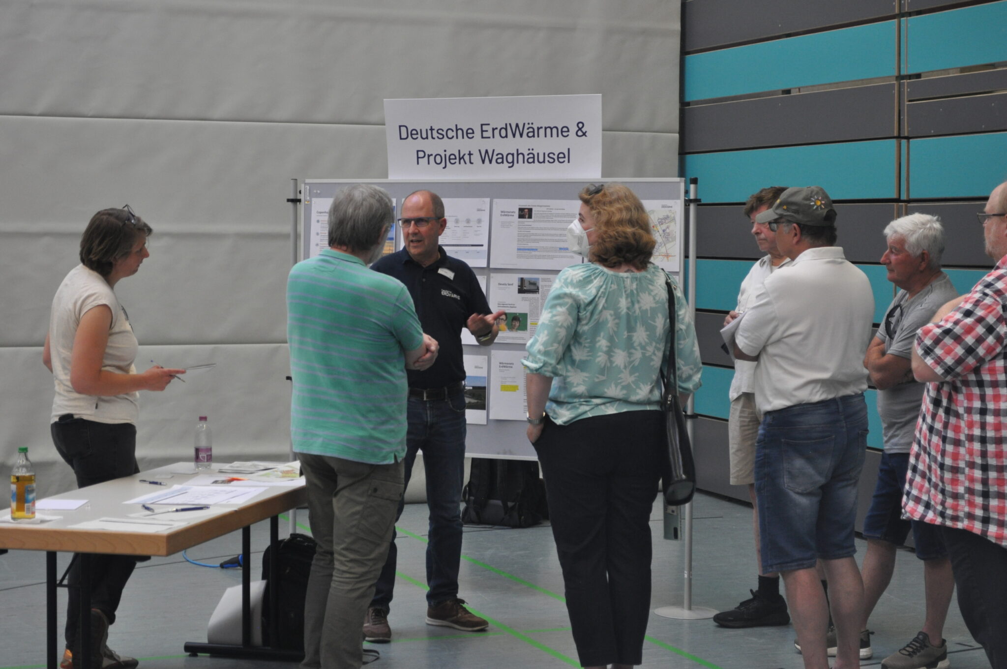 Eine Gruppe von Menschen steht vor einem Informationsdisplay mit der Aufschrift Deutsche ErdWärme & Projekt Waghäusel in einer Sporthalle und informiert sich über Tiefengeothermie, während ein Referent spricht. In der Nähe stehen Tische mit Unterlagen und Getränken.