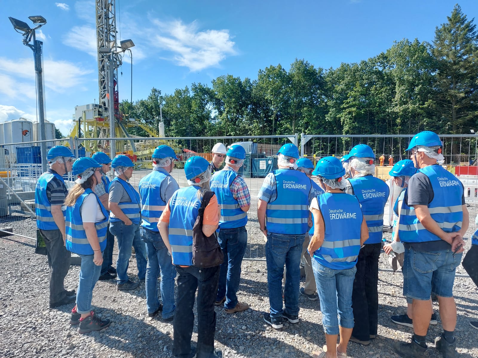 Eine Gruppe von Personen mit blauen Helmen, Sicherheitswesten und Gesichtsmasken steht auf einer Baustelle für Tiefengeothermie- oder Wärmeversorgungsprojekte im Freien zusammen, umgeben von einem Zaun, mit Bäumen und Geräten im Hintergrund.