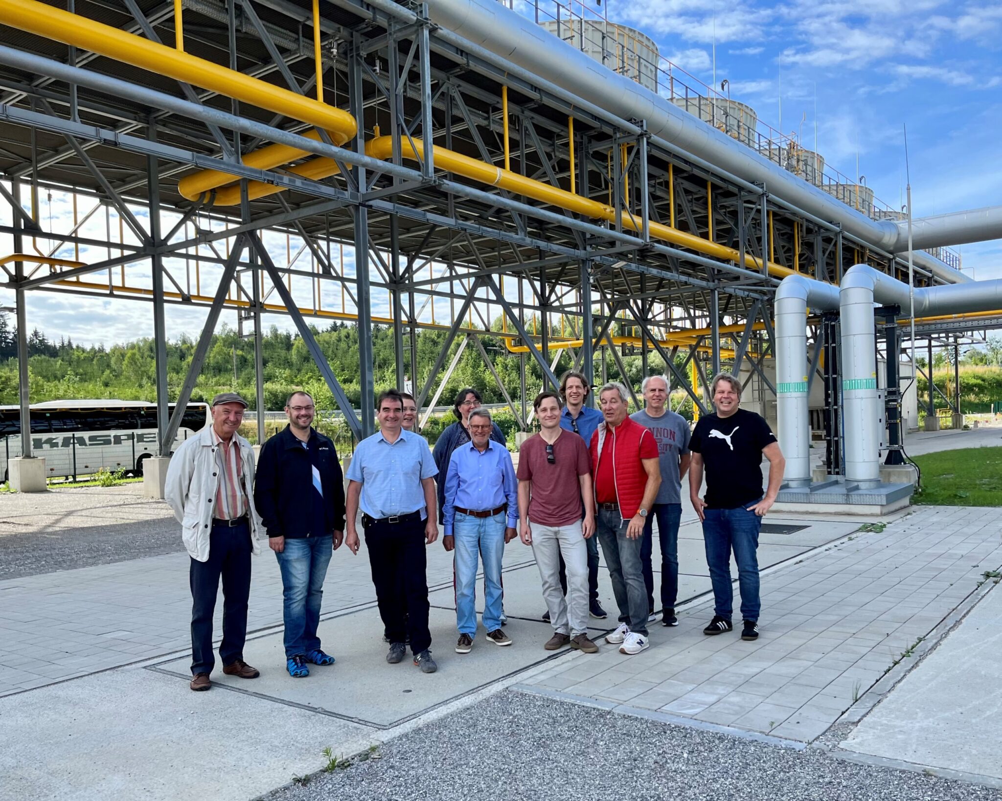 Eine Gruppe von elf Personen steht lächelnd vor einer Industrieanlage mit großen Metallrohren und -strukturen, die auf die Bemühungen hinweisen, Wärmenetze für die Dekarbonisierung auszubauen, umgeben von Grün und einem blauen Himmel.