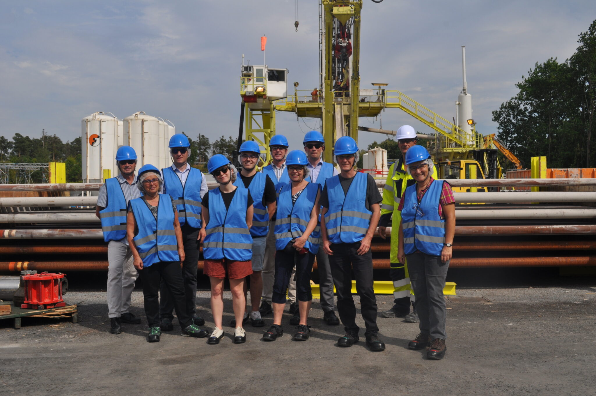 Eine Gruppe von Personen mit blauen Sicherheitswesten und blauen Helmen steht auf einem Industriegelände mit Schwerpunkt Tiefengeothermie zusammen. Im Hintergrund sind Rohre und Bohrausrüstung zu sehen, die die nachhaltige Wärmeversorgung unterstützen.