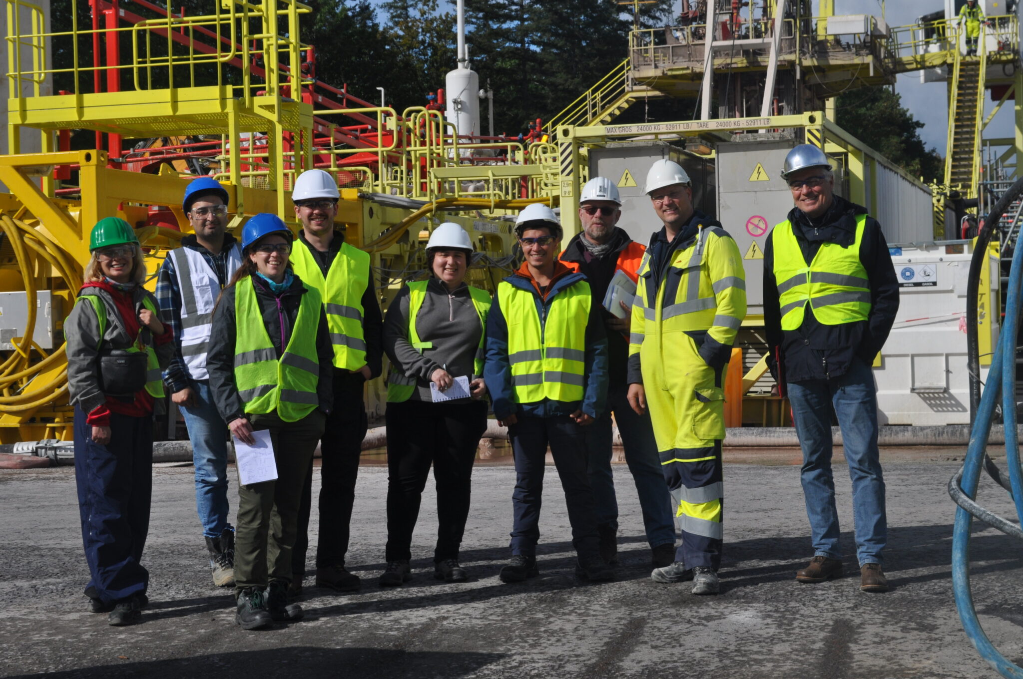 Eine Gruppe von Personen mit Sicherheitswesten und Schutzhelmen steht im Freien auf einem Industriegelände mit gelben Metallstrukturen zusammen und unterstützt die Dekarbonisierung und den Ausbau von Wärmenetzen im Bereich der Wärmeversorgung.