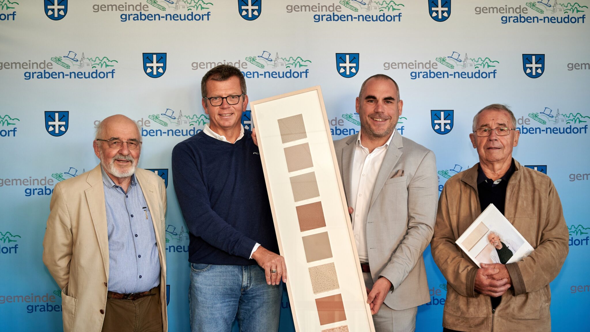 Vier Männer stehen vor einer Kulisse der Gemeinde Graben-Neudorf; zwei halten einen vertikalen Rahmen mit Farbmustern, einer ein Porträtfoto. Alle lächeln in legerer Business-Kleidung bei einer Veranstaltung der Deutschen Erdwärme Tiefengeothermie.