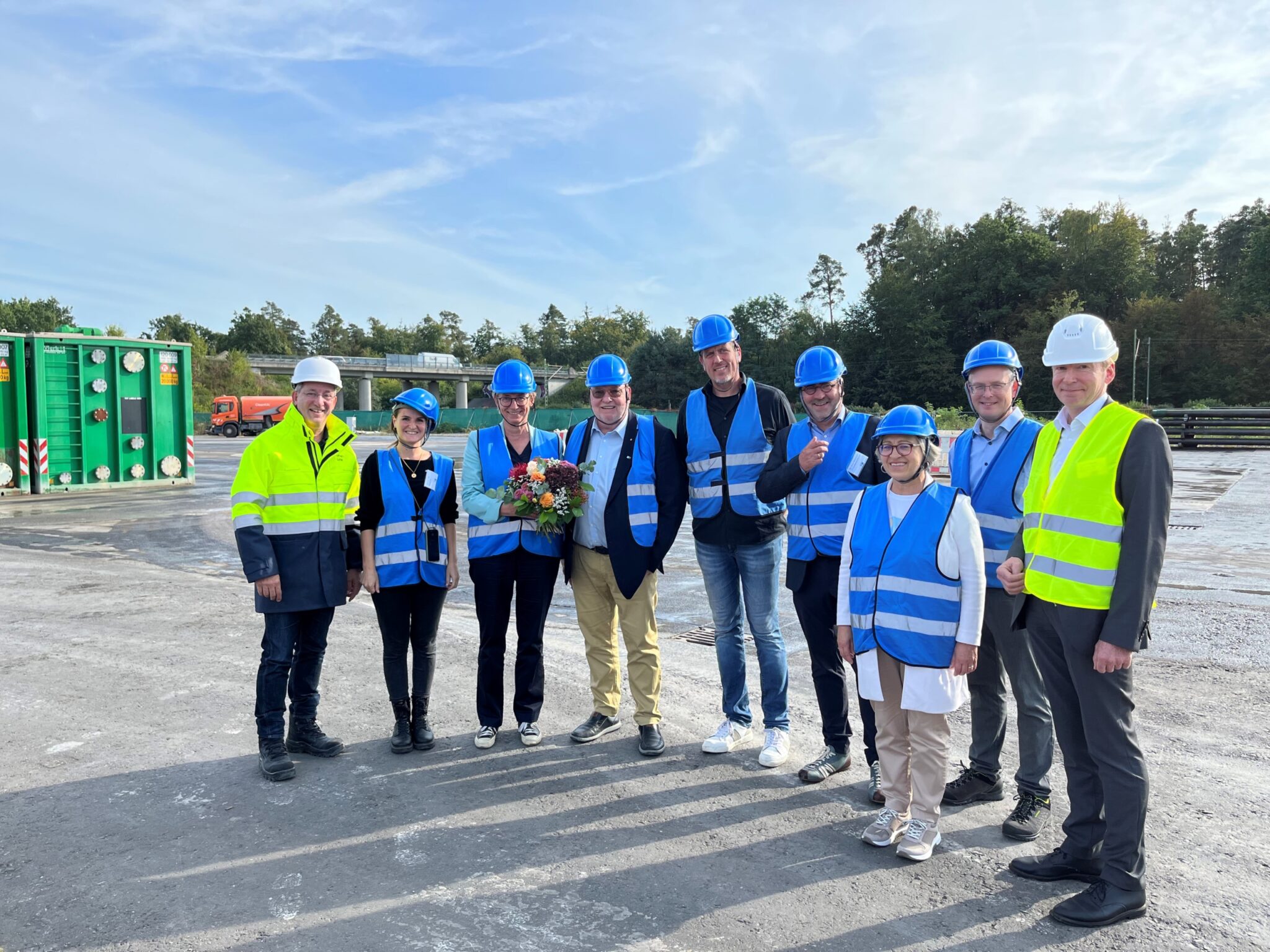 Besuchergruppe der IHK mit Schutzhelmen und Warnwesten am Geothermiebohrplatz der Deutschen Erdwärme in Graben-Neudorf. Eine Frau hält einen Blumenstrauß in der Hand, da sie die 2000 Besucherin am Bohrplatz ist.