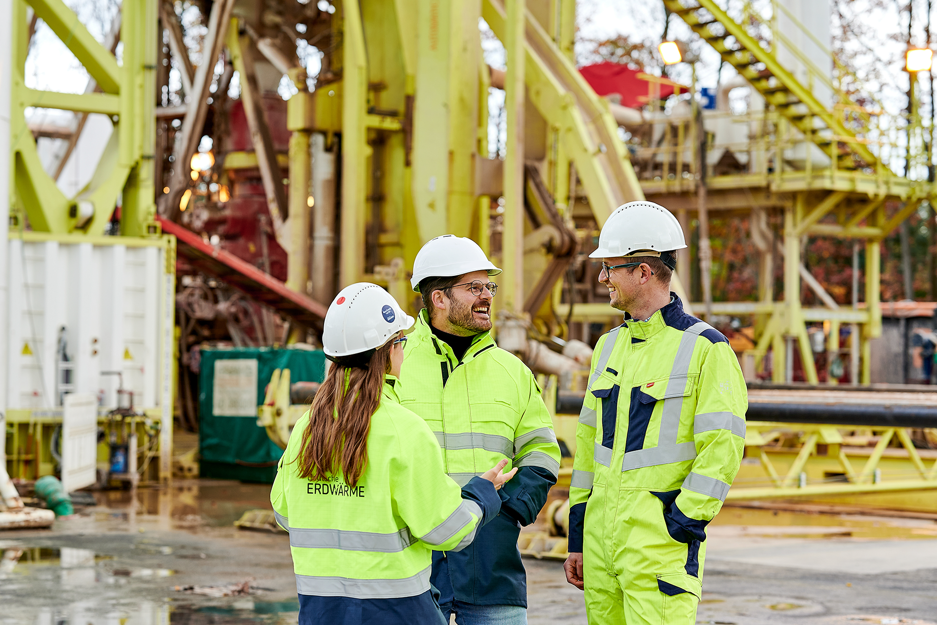 Drei Arbeiter in gelben Sicherheitswesten und weißen Helmen stehen zusammen, lächeln und unterhalten sich auf einem Industriegelände, das auf Dekarbonisierung ausgerichtet ist, mit großen Maschinen und Metallstrukturen im Hintergrund.