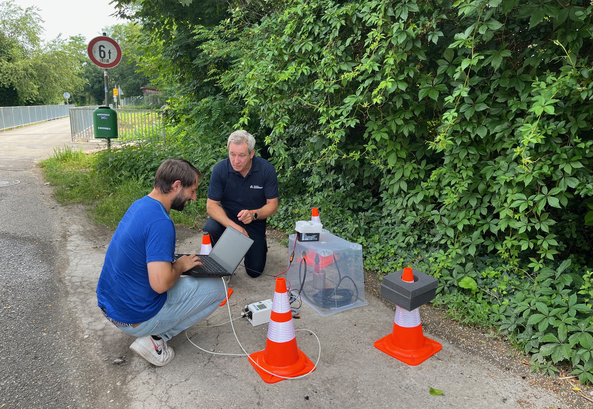 Zwei Männer knien am Straßenrand, benutzen einen Laptop und Überwachungsgeräte, die zwischen Verkehrsleitkegeln neben dichten grünen Büschen aufgestellt sind, während sie Tiefengeothermie-Messungen für die deutsche Erdwärme auf einer ruhigen Straße durchführen.