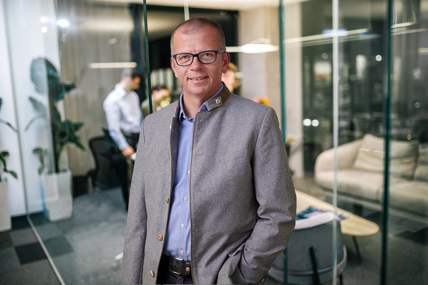 Ein Mann mit Brille, blauem Hemd und grauem Blazer steht lächelnd in einem modernen Büro mit Glaswänden - eine ideale Umgebung für Teams, die sich auf Dekarbonisierung konzentrieren, während andere im Hintergrund zusammenarbeiten.