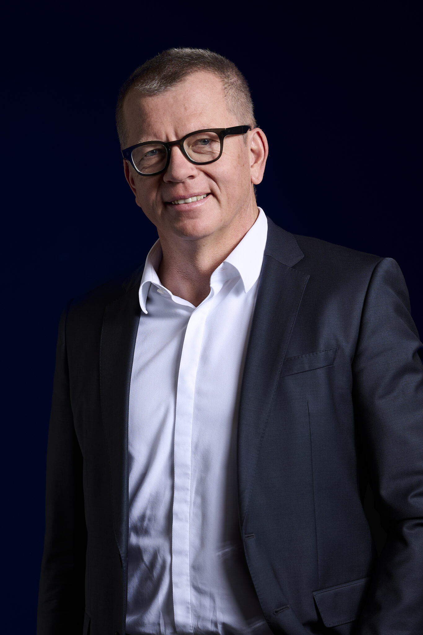 Ein Mann mit Brille, weißem Hemd und dunkler Anzugsjacke steht vor einem dunklen Hintergrund und lächelt leicht in die Kamera - ein Hinweis auf die Professionalität, mit der deutsche Erdwärme zu Wärmenetzen für eine zuverlässige Wärmeversorgung ausgebaut wird.