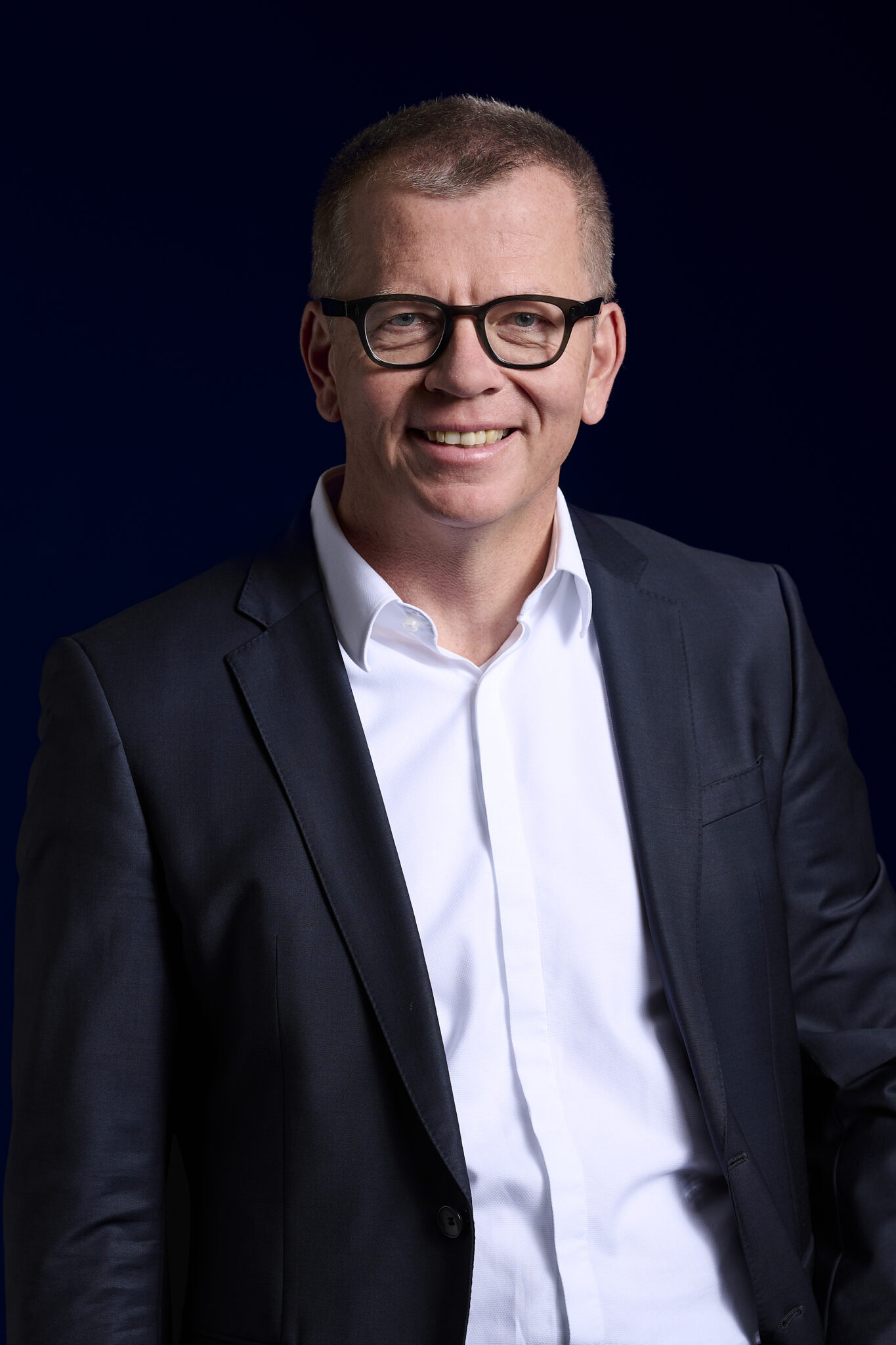 Ein lächelnder Mann mit kurzen Haaren und Brille trägt eine dunkle Anzugsjacke über einem weißen Hemd und steht vor einem dunkelblauen Hintergrund, der den innovativen Geist der deutschen Erdwärme in nachhaltiger Wärmeversorgung widerspiegelt.