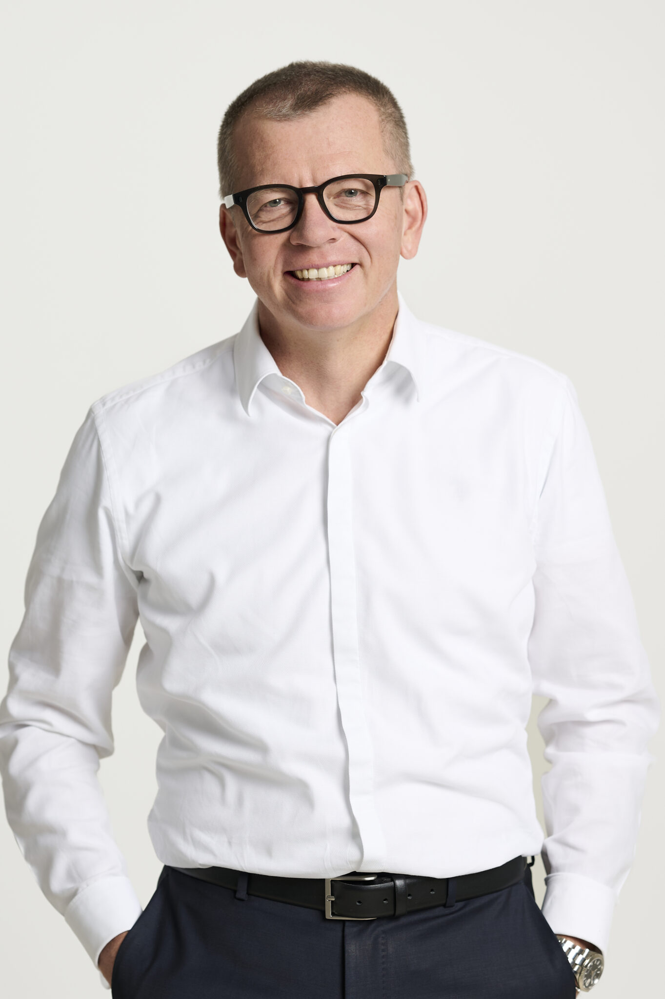 Ein lächelnder Mann mit Brille, weißem Button-Down-Hemd und dunkler Hose steht vor einem schlichten hellen Hintergrund, eine Hand in der Tasche, und verkörpert den innovativen Geist der deutschen Erdwärme und das Engagement für eine nachhaltige Wärmeversorgung.